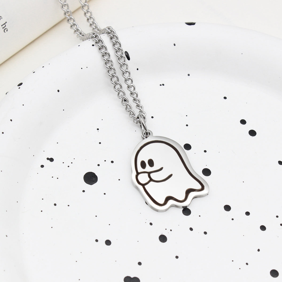 Pendentif fantôme timide sur chaîne en acier, posé sur une assiette blanche mouchetée