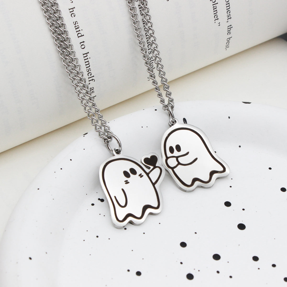 Deux pendentifs fantôme sur chaîne en acier, posés sur une assiette et un livre ouvert